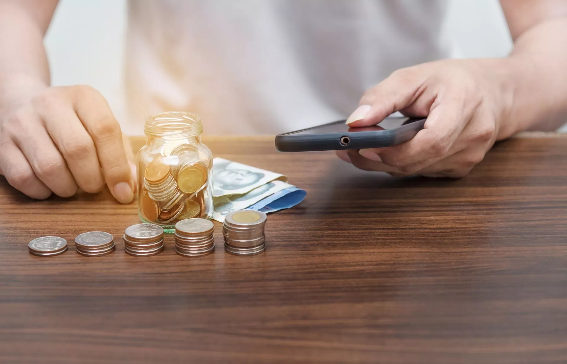 桌面上擺放零錢與紙鈔，一手將硬幣放入玻璃罐中，另一手拿著手機，呈現以數位工具進行理財與儲蓄的情境。