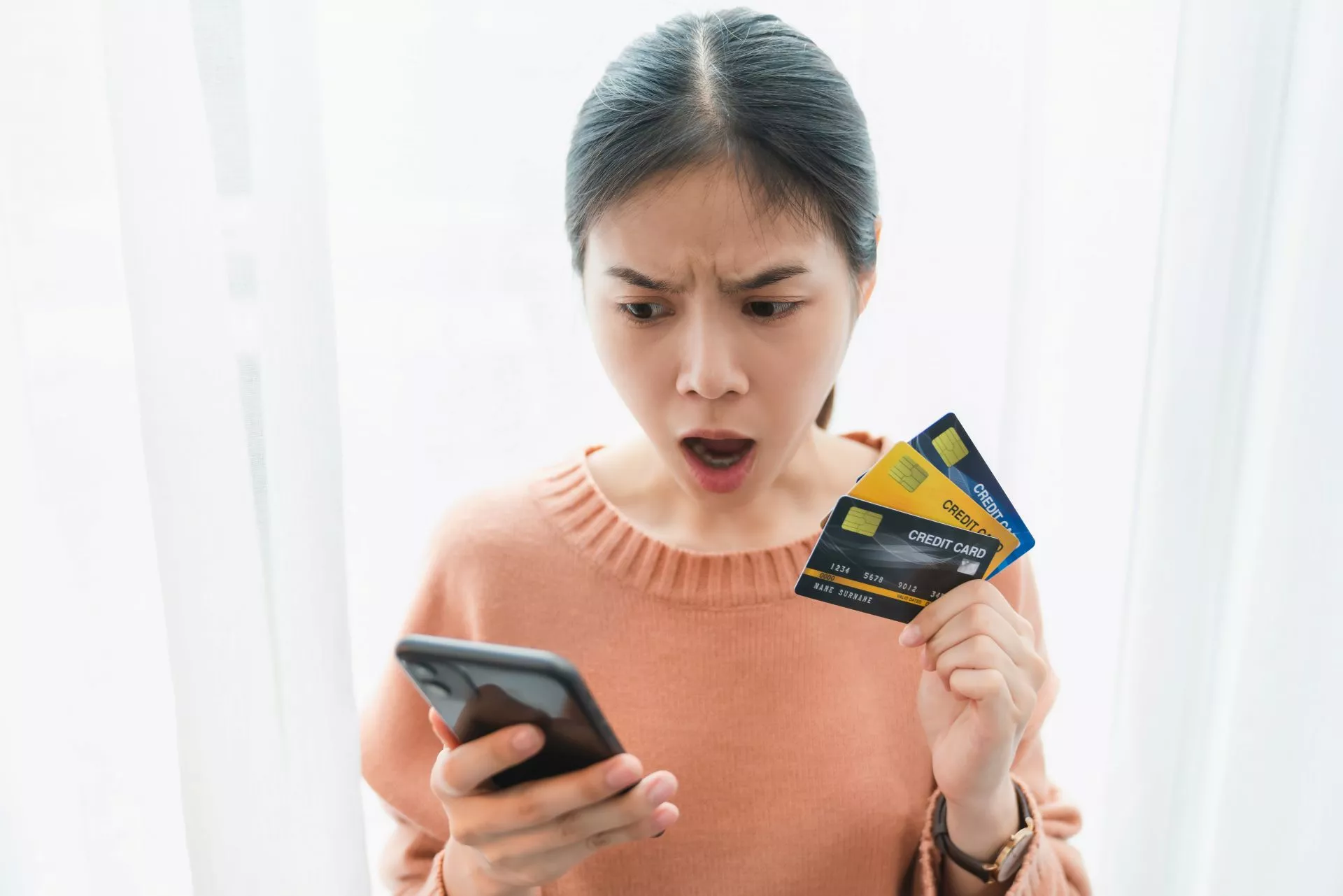 一名女子手持多張信用卡並盯著手機螢幕，表情驚訝，呈現查看帳務或付款異常的情境。