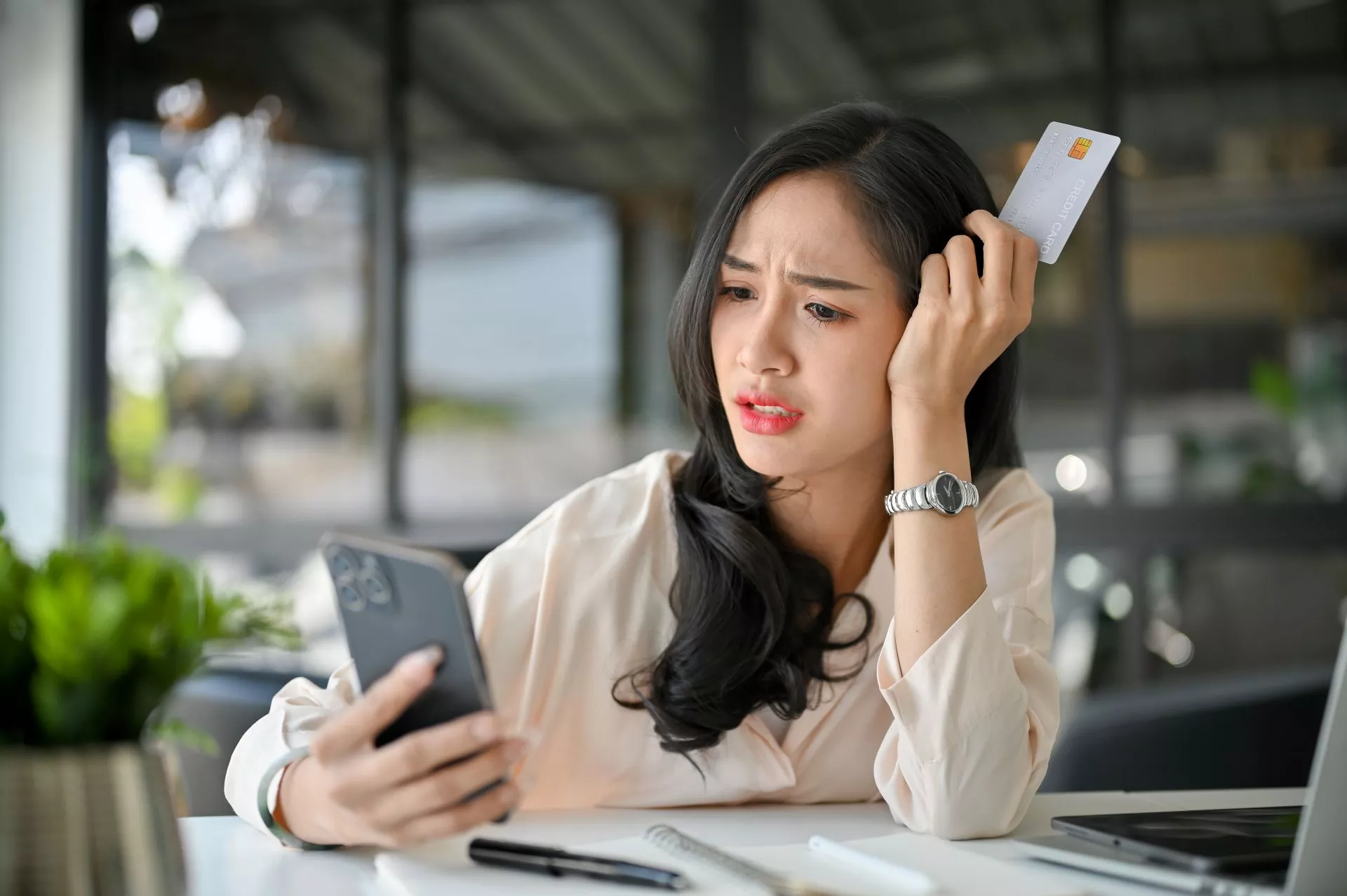 女性坐在室內桌前，一手拿著手機查看訊息，另一手拿著信用卡靠在頭旁，表情焦慮，桌上有筆記本與筆電，呈現消費或付款困擾情境。